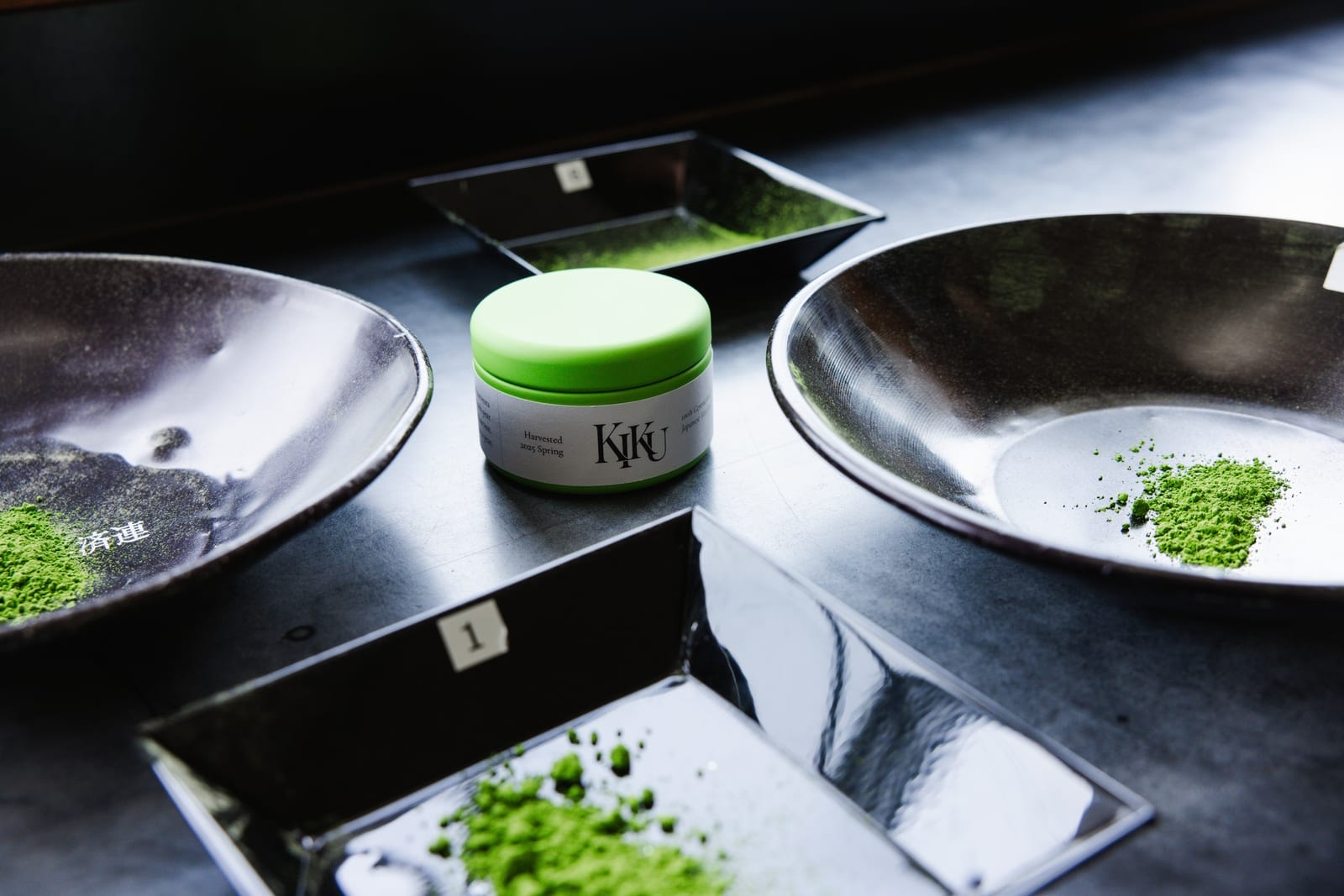 Green RIKYU matcha tin between black bowls of matcha powder