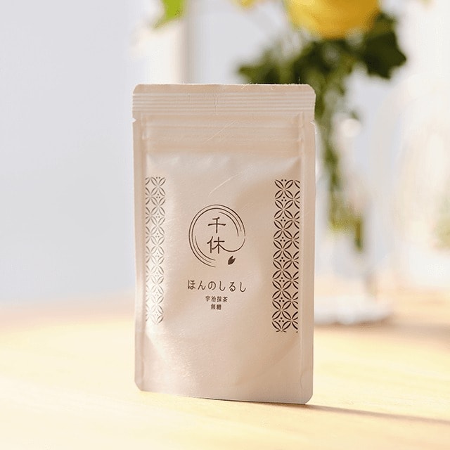 A white Senkyu zip pouch of Uji matcha standing on a wooden table in front of flowers